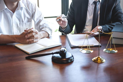 A divorce lawyer speaks to a client while both sit in front of a table with a gavel and scales. 